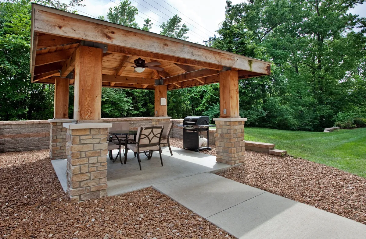 A welcoming outdoor area featuring a wooden gazebo supported by stone pillars, an outdoor dining table, and a grill. The space is surrounded by greenery, creating a relaxed setting ideal for family meals and social gatherings.