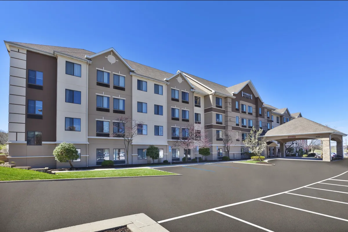 A welcoming exterior shot of the hotel entrance featuring large windows, an inviting awning, manicured landscaping, and a glimpse of the lobby inside. The scene suggests accessibility and proximity to local destinations, setting a positive first impression for business travelers.