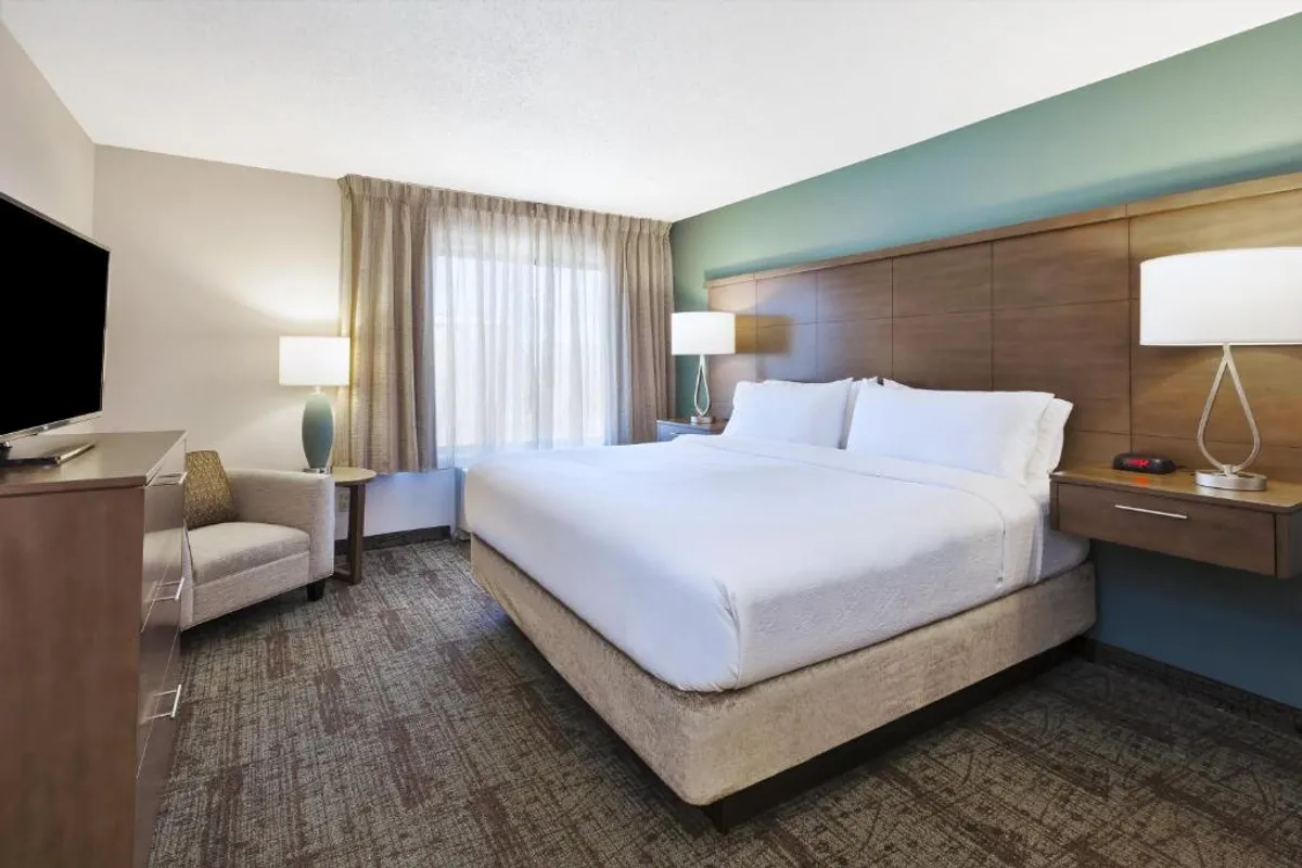 A serene bedroom retreat with a plush king bed dressed in crisp, high-quality linens. The room features soft lighting, a seating area near a large window with blackout curtains, nightstands with reading lamps, and a mounted TV for relaxation. The overall mood is calm and inviting, perfect for winding down after a busy day.
