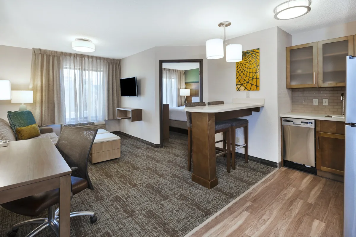 The image showcases a bright, welcoming living space with a plush seating area, a modern kitchenette, a cozy dining counter, and tasteful artwork. The area is designed for both relaxation and productivity, featuring comfortable furnishings and practical amenities.